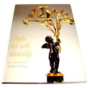 May include: A hardcover book with a white cover featuring a dark bronze cherub statue holding a golden floral arrangement. The text on the cover reads: Objets des arts décoratifs, Les collections privées de Saint-Pétersbourg in gold lettering.