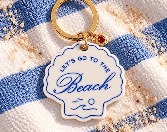 May include: A white shell-shaped keychain with a gold-tone metal ring and a small red crystal charm. The keychain features blue text that reads "LET'S GO TO THE BEACH" and a simple blue graphic of a sun, waves, and birds.