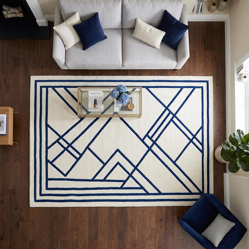 Pode incluir: Um tapete retangular com uma base de cor creme e um padr&atilde;o geom&eacute;trico azul-escuro arrojado, composto por linhas que se cruzam e formas angulares. O design inclui uma moldura espessa e v&aacute;rias linhas diagonais na superf&iacute;cie.
