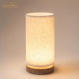 May include: A cylindrical table lamp featuring a textured off-white fabric shade that emits a warm glow. The lamp is supported by a circular wooden base with a natural grain finish. The text LumenCraft Co. LIGHT THAT SETS THE MOOD is visible.