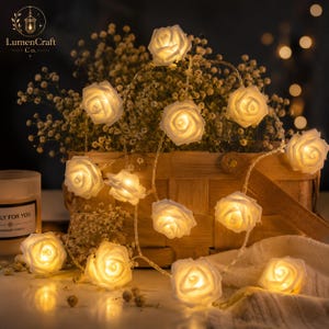 Puede incluir: Una cadena de luces LED con múltiples cubiertas en forma de rosa blanca que emiten un brillo cálido. Las rosas están dispuestas en un grupo decorativo, con flores individuales iluminadas desde el interior para un efecto suave.