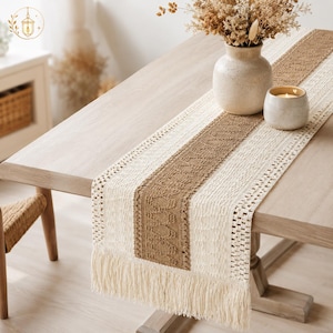 May include: A rectangular table runner featuring a central brown woven panel flanked by cream-coloured textured sections with open-weave borders and long fringe at the ends. The runner is placed on a light wood table with a ceramic vase and candle.