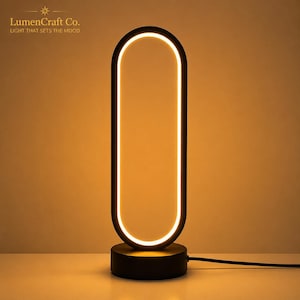 以下が含まれることがあります： 内側の縁に沿って連続した統合型LEDライトストリップを備えた、黒い楕円形のテーブルランプ。ランプは円形のベースを特徴とし、暖かい光を放ちます。左上隅にLumenCraft Co. LIGHT THAT SETS THE MOODというテキストが表示されています。