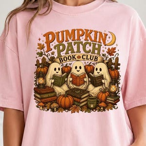 May include: A light pink t-shirt featuring a graphic print with the text "PUMPKIN PATCH BOOK CLUB". The design shows three ghosts reading books surrounded by pumpkins, lanterns, and autumn leaves in shades of orange, brown, and green.