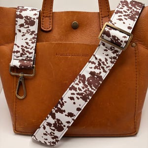 May include: A brown leather tote bag with a detachable strap featuring a white and brown cowhide pattern. The bag has a front pocket and top handles. The strap is adjustable and attaches with a metal clasp.