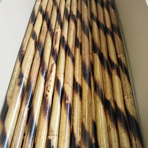 May include: A collection of cylindrical bamboo poles arranged in parallel. Each pole features a natural light tan surface with dark brown, charred diagonal stripes wrapping around the length of the wood, creating a consistent pattern across the group.
