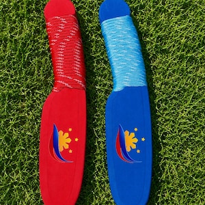 May include: Two foam training knives, one red and one blue, each featuring a handle wrapped in matching paracord. The blades display a yellow sun and three stars emblem with a curved red and blue accent graphic printed on the flat surface.