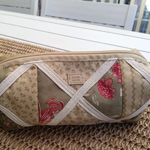 May include: A rectangular fabric pouch featuring a patchwork design in shades of olive green, beige, and pink floral patterns. The exterior is accented with white woven ribbon strips arranged in a crisscross pattern and a small rectangular label reading HAND MADE.