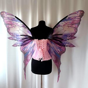 Puede incluir: Un corsé rosa claro con cierre de cordones en la espalda combinado con grandes alas de hada translúcidas. Las alas presentan un patrón degradado detallado en tonos morado, magenta y azul con estructuras de venas oscuras y definidas.