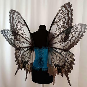 May include: A bright teal corset with a lace-up back closure, paired with large, translucent black butterfly wings featuring intricate, dark, vein-like patterns and scalloped edges, displayed on a black mannequin torso against a white background.