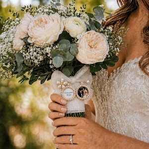 Könnte beinhalten: Ein Blumenstrauß mit blassrosa und weißen Gartenrosen, Schleierkraut und Eukalyptusblättern. Die Stiele sind in weißen Stoff gewickelt, an dem zwei runde Fotoanhänger befestigt sind, einer mit einem Paar und der andere mit Text.