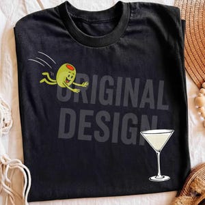 Könnte beinhalten: Ein schwarzes Kurzarm-T-Shirt mit einer grünen Cartoon-Olive mit Armen und Beinen, die auf ein Martiniglas zuspringt. Der Text ORIGINAL DESIGN ist in einer großen, dunkelgrauen Schrift mittig auf dem T-Shirt hinter der Grafik aufgedruckt.