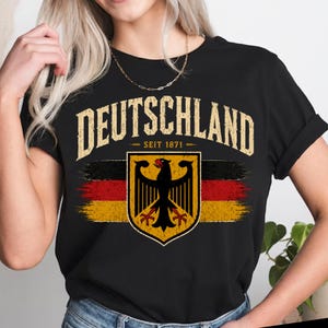 May include: A black t-shirt featuring the text DEUTSCHLAND SEIT 1871 in a distressed font above a graphic of the German eagle emblem centered over a horizontal brushstroke design in the colors of the German flag: black, red, and gold.