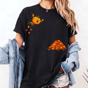 Puede incluir: Una camiseta negra de manga corta con un gráfico de una calabaza naranja sonriente saltando hacia un montón de hojas de otoño coloridas. La calabaza está situada en la parte superior izquierda, mientras que el montón de hojas rojas, naranjas y amarillas se encuentra en la parte inferior derecha.