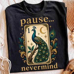May include: A black crew neck t-shirt featuring a graphic of a peacock perched on a branch inside a rectangular frame. The text "pause..." appears above the frame and "nevermind" below. The design includes gold stars, a crescent moon, and peacock feathers.