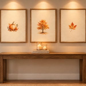 Puede incluir: Tres cuadros de pared enmarcados con ilustraciones en acuarela de una calabaza, un árbol de hoja caduca con follaje naranja y una hoja de arce. Las láminas están en marcos de madera clara sobre una mesa consola de madera con velas.