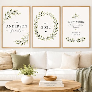 May include: Three vertical wall art prints in light wood frames. The left print features green leaves and the text THE ANDERSON family. The centre print shows a green leaf wreath and the text EST. 2022. The right print displays NEW YORK, coordinates, and leaves.