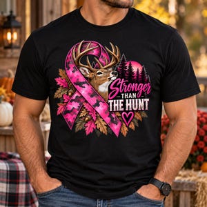 May include: A black t-shirt featuring a graphic of a deer head inside a pink camouflage awareness ribbon. The design includes autumn leaves, a forest silhouette, and the text "Stronger THAN THE HUNT" with a small pink heart icon below it.