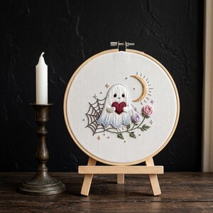 Könnte beinhalten: Ein Stickrahmen mit einem weißen Geist, der ein rotes Herz hält, umgeben von einem Spinnennetz, einem goldenen Halbmond und kleinen rosa und violetten Rosen auf weißem Stoff, präsentiert auf einer kleinen Holzstaffelei neben einer weißen Kerze.