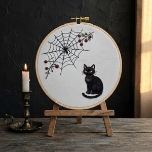 Könnte beinhalten: Ein runder Stickrahmen aus Holz mit weißem Stoff, auf dem eine schwarze Katze, ein Spinnennetz und kleine rote Beeren gestickt sind. Der Rahmen steht auf einer kleinen Holzstaffelei neben einer weißen Kerze in einem Messingständer.