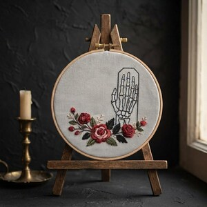 Könnte beinhalten: Ein runder Stickrahmen mit hellgrauem Stoff, der den schwarzen Umriss einer Skeletthand und ein gesticktes Arrangement aus roten Rosen mit grünen Blättern zeigt. Der Rahmen steht auf einer kleinen hölzernen Staffelei.
