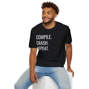 May include: A black short-sleeved t-shirt featuring the white text "COMPILE. CRASH. REPEAT." printed on the front. The shirt has a crew neckline and a standard fit, providing a clean and simple look for everyday wear in a casual setting.
