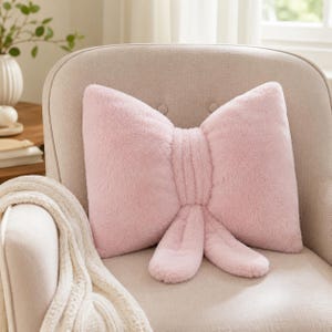 Peut inclure: Un coussin en peluche rose clair doux en forme de grand nœud. Le coussin présente une surface texturée et duveteuse avec un nœud central et deux boucles pendantes à la base, imitant l'apparence d'un ruban noué dans un coloris rose pastel uni.