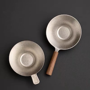 Puede incluir: Dos coladores de té circulares de metal con un acabado plateado mate y texturizado. Cada colador presenta una zona central de malla perforada para filtrar. Uno tiene un mango corto de metal, mientras que el otro tiene un mango largo de madera.