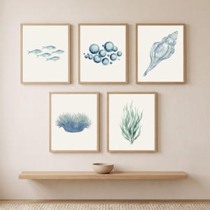 May include: A set of five framed wall art prints featuring watercolor illustrations of marine life and ocean elements, including a school of fish, blue bubbles, a seashell, a sea anemone, and green seaweed, all displayed in light wood frames.