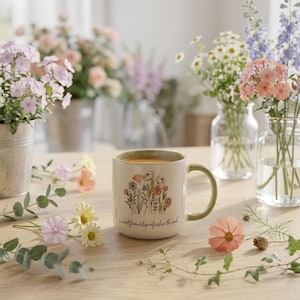 May include: A cream-colored ceramic mug with a green handle and rim, featuring a colorful floral illustration and the text 'a sweet friendship refreshes the soul' printed on the front, sitting on a wooden surface surrounded by loose flowers.