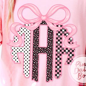 May include: A light pink t-shirt featuring a large central graphic of a pink bow above a three-letter monogram. The letters are filled with black and white polka dots, a black and white heart pattern, and a black and white checkerboard print.