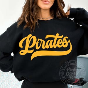 May include: A black long-sleeve crewneck sweatshirt featuring the word Pirates printed in a large, bold, yellow cursive font across the chest. The design has a prominent underline flourish extending from the final letter of the word.