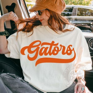 May include: A cream-colored short-sleeve t-shirt featuring the word Gators printed in a large, bold, orange cursive font across the chest. The shirt has a crew neckline and a relaxed fit, with a watermark logo visible in the lower right corner.