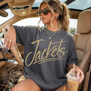 Op de afbeelding: Een antracietgrijs T-shirt met korte mouwen en het woord Jackets in een groot, cursief goudkleurig lettertype op de borst. Het shirt heeft een ronde hals en een relaxte pasvorm, gedragen met een gelaagde gouden ketting en gouden ringen aan de vingers.