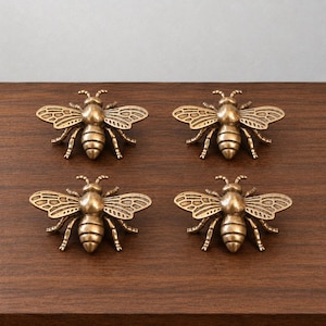May include: A set of four identical bee-shaped drawer knobs made of a metallic material with a bronze-toned finish. Each knob features detailed wings with etched lines, a rounded thorax, and a segmented abdomen, designed for furniture hardware.