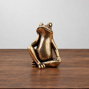 May include: A bronze-colored frog figurine in a seated, contemplative pose. The frog has a textured, metallic finish with dark patina accents in the crevices, resting one hand against its cheek while looking upward with prominent eyes.