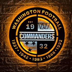 May include: A circular black metal wall sign featuring the Washington Football Commanders logo with backlighting. The text reads WASHINGTON FOOTBALL, COMMANDERS, EST 1932, and includes the years 1937, 1942, 1983, 1988, 1992, and three stars.