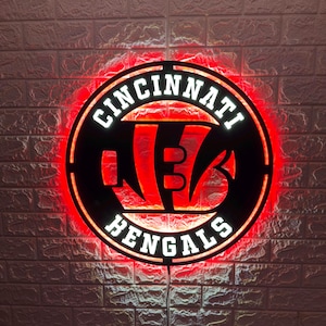 Può includere: Un'insegna circolare da parete con il logo dei Cincinnati Bengals in nero con scritte bianche e una vivace retroilluminazione a LED rossa. Il design include il nome della squadra in grassetto attorno all'emblema centrale a forma di B.