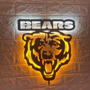 Può includere: Un'insegna a LED da parete con la parola BEARS in lettere maiuscole illuminate di bianco, posizionata sopra una sagoma stilizzata di testa d'orso con contorno nero che emana una calda luce giallo oro dal retro del design.