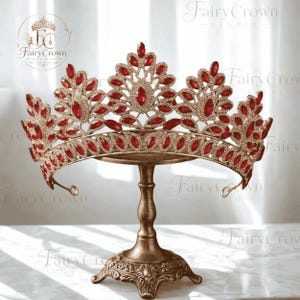 Op de afbeelding: Een goudkleurige kroon met een symmetrische opstelling van marquise-geslepen rode kristallen en kleine heldere strass-steentjes. De kroon heeft een hoge centrale piek en twee kleinere zijpieken, allemaal versierd met rode stenen in een bladvormig patroon.