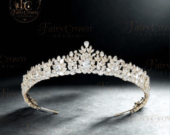 Könnte beinhalten: Ein goldfarbenes Diadem mit einer dichten Anordnung aus klaren, marquise- und birnenförmigen Kristallen. Das Kopfstück zeigt ein symmetrisches, spitz zulaufendes Design mit einem zentralen birnenförmigen Stein an der Spitze auf einem dünnen Metallreif.