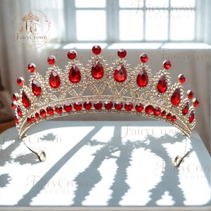 May include: A gold-toned metal tiara featuring a series of teardrop-shaped red crystals and smaller round red stones. The frame is encrusted with numerous tiny clear rhinestones arranged in a decorative pattern, with a curved base and two loops.