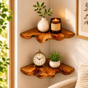 May include: Two stacked corner shelves made of natural wood with live edges. The top shelf holds a white ceramic vase with green leaves and a brown glass candle jar labeled Hinoki Fog. The bottom shelf holds a gold analog clock and a small succulent.