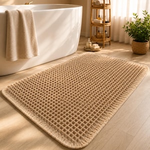 May include: A rectangular beige bath mat featuring a raised grid or waffle-like texture across its surface. The mat has rounded corners and a thick, woven border that frames the central square pattern, providing a consistent tan color throughout.
