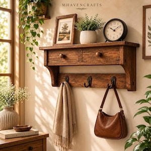 May include: A wall-mounted wooden shelf featuring two drawers with round knobs and three black metal hooks underneath. The shelf holds a framed landscape print, a potted green plant in a ribbed white container, and a black analog clock.