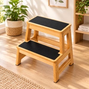 May include: A two-step stool constructed from light-toned bamboo wood, featuring two rectangular platforms with black non-slip grip tape surfaces. The stool has a sturdy frame with four legs and horizontal support bars connecting the sides.