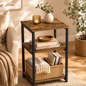 May include: A three-tier side table with a dark brown wood-grain finish and a black metal frame. The top shelf holds a ribbed glass candle holder and a white vase. The middle shelf contains two stacked books and a small wooden bowl.