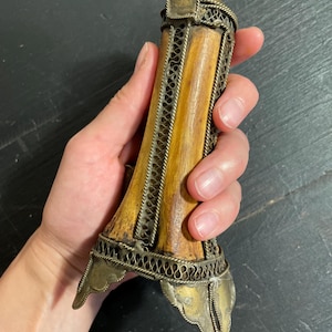 Peut inclure: Un objet cylindrique doté d'un noyau central en bois brun clair, entouré d'un filigrane métallique argenté complexe. La pièce est surmontée de plaques métalliques décoratives aux deux extrémités, la base présentant trois extensions pointues.