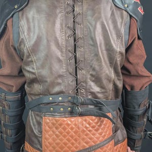 Op de afbeelding: Een bruine en beige jas met een vetersluiting op de rug met metalen oogjes, een gewatteerd onderste paneel en zwarte lederen schouderstukken. Het kledingstuk bevat zwarte gespriemen bij de taille en onderarmen, met rode accenten op de schouders.