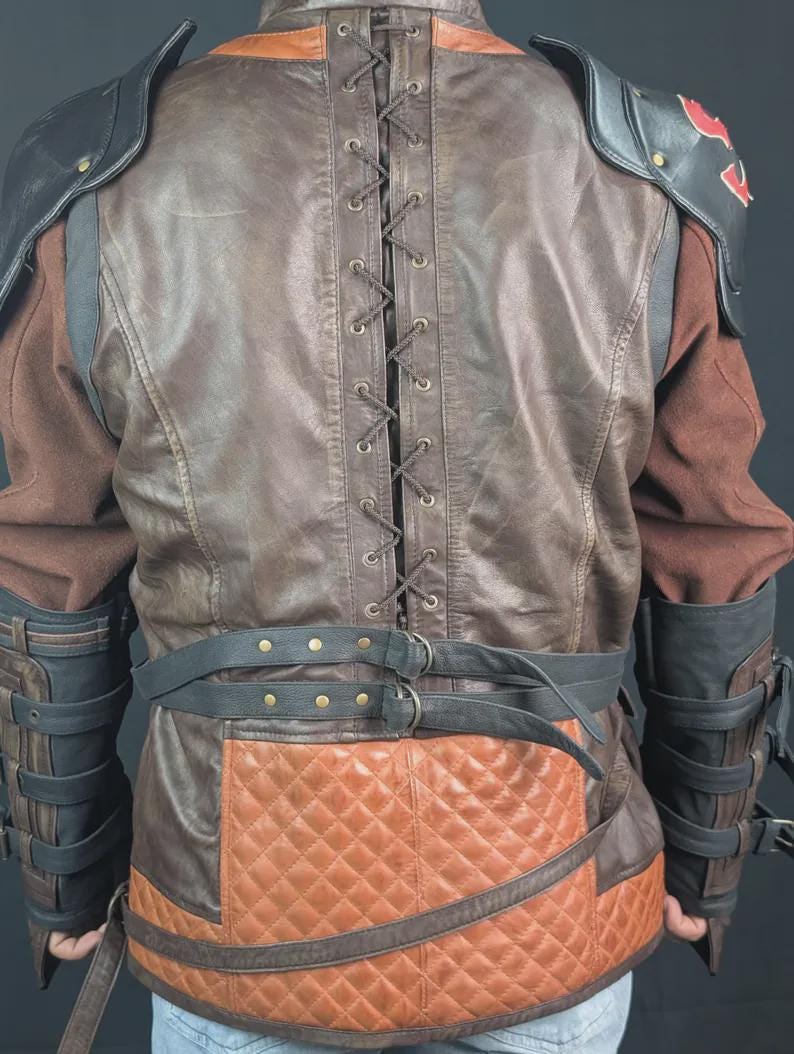 Op de afbeelding: Een bruine en beige jas met een vetersluiting op de rug met metalen oogjes, een gewatteerd onderste paneel en zwarte lederen schouderstukken. Het kledingstuk bevat zwarte gespriemen bij de taille en onderarmen, met rode accenten op de schouders.